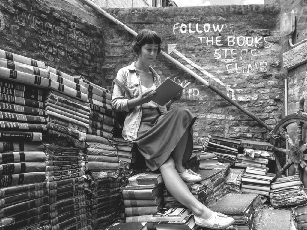 Eine Frau sitzt auf Stapeln alter Bücher, ein Buch lesend.
